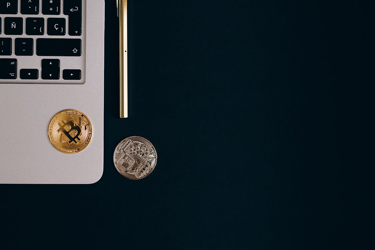 Close-up view of Bitcoin and Ethereum coins next to a laptop and pen on a black surface.